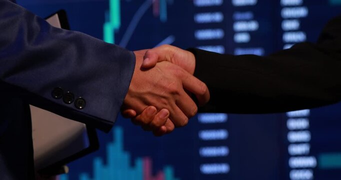 Close-up of a business handshake in front of a blurred market chart screen, symbolizing deal, trust, partnership, and finance success