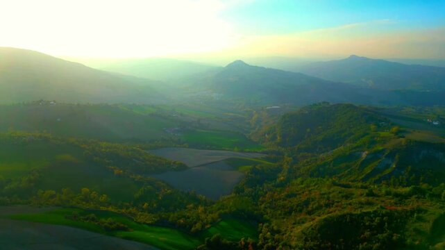 Mystical aerial pullback truck right pan right shot near Santa Vittoria in Matenano, hazy misty Sibillini Mountains in the distance, imbued with spiritual sunlight, ravines and hills in the foreground