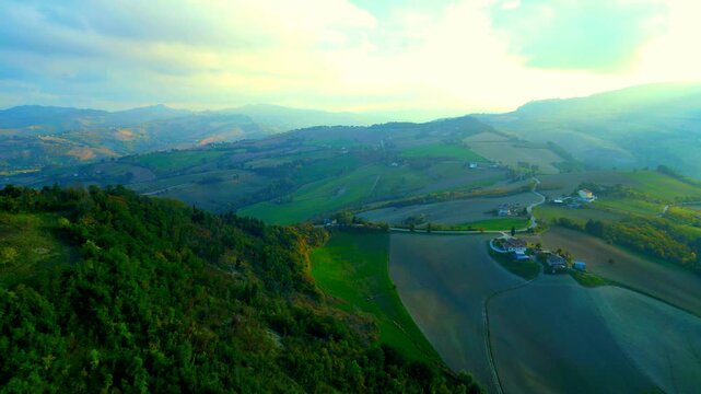 Soothing aerial pedestal up push-in truck right pan left shot near Santa Vittoria in Matenano, tiny roads stretching atop lofty hillscapes, cars passing by, hazy sunlight imbuing the view 
