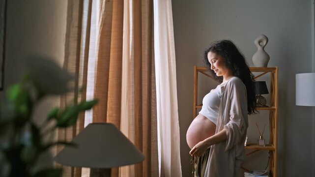 Pregnant woman opening curtains morning light. Expecting mother cradling belly