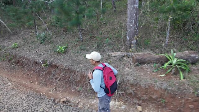 Slow motion side view of a man wearing a cap and red backpack running along a rocky path in a green forest. Active outdoor adventure, hiking, and nature lifestyle concept.