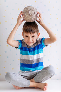 A cheerful young boy in a striped polo balances a textured papier‑m&acirc;ch&eacute; moon atop his head. He sits cross‑legged on a white surface against a star‑patterned backdrop, exuding playful curiosity.