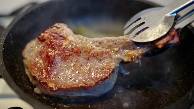 Chef using metal tongs to flip a raw pork chop while searing it in a hot iron skillet with aromatic rosemary and fresh garlic cloves.