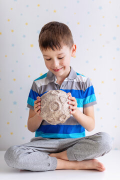 A cheerful young boy in a striped polo holds a textured papier‑m&acirc;ch&eacute; moon with realistic craters. He sits cross‑legged on a white surface against a star‑patterned backdrop, exuding playful curiosity.