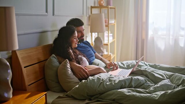 Expecting couple browsing laptop lying bed. Pregnant wife husband online