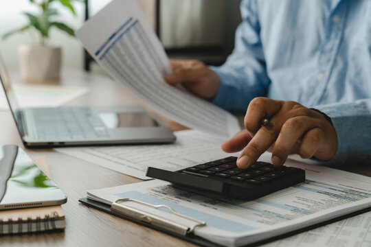man use calculator while holding a financial document beside a laptop, showing budgeting, accounting, and report review workflow for corporate finance planning process.