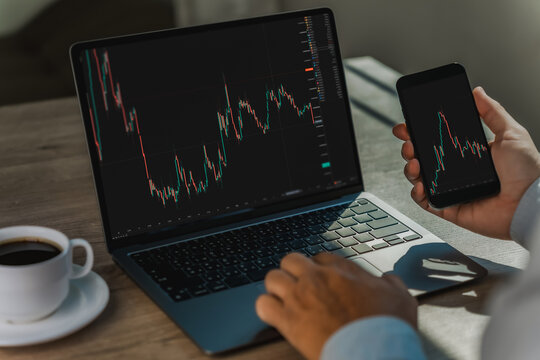 Business person reviewing stock market data on laptop and smartphone, comparing candlestick charts across digital screens for trading analysis, investment research, market monitoring.