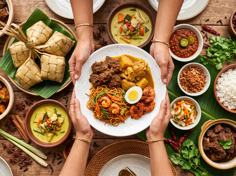 Top view of a festive malay communal feast with hands holding a plate of noodles, beef rendang, and chicken curry on a wooden table