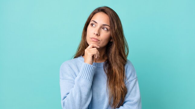 Thoughtful young woman looking up while thinking deeply against a solid turquoise blue studio background