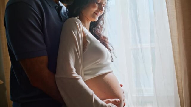Pregnant woman husband embrace home. Expecting couple holding belly by window.
