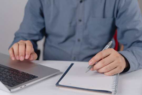 Man typing on laptop keyboard and holding pen over blank notebook at desk