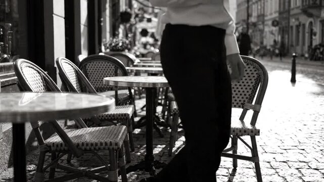 Waiter walks past cafe tables with outdoor wicker chairs along cobblestone street sidewalk during morning daylight city scene in black and white video footage.
