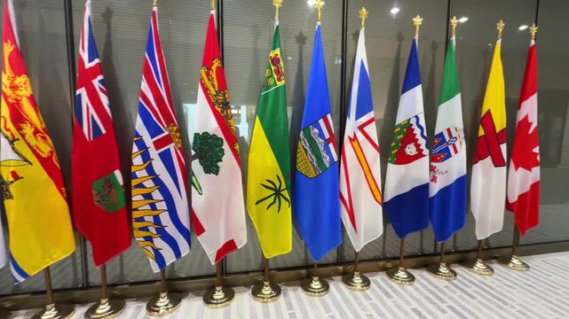 Wide shot of Canadian national, provincial, and territorial flags displayed indoors on gold brass stands in Ottawa, ON, Canada. 