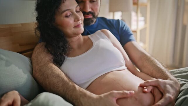 Pregnant belly closeup, husband hands caressing wife. Expecting parents on bed.