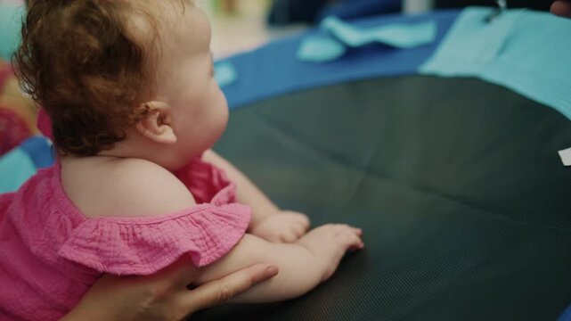 Infant girl in a pink dress learning to bounce on a trampoline with adult supervision during outdoor playtime