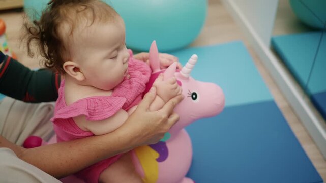 Baby girl receiving support from therapist, sitting on a pink unicorn toy during a physical therapy session for motor skills development