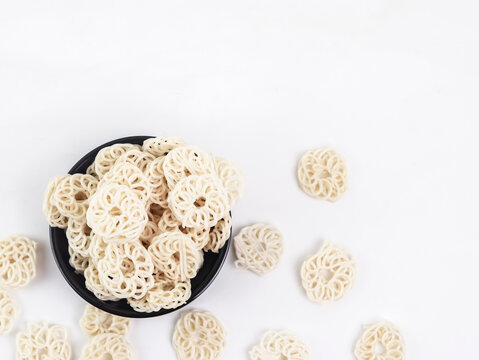 Top-down view of a black bowl filled with raw white kerupuk udang (Indonesian prawn crackers or tapioca crackers) with loose crackers scattered around on a clean white background.