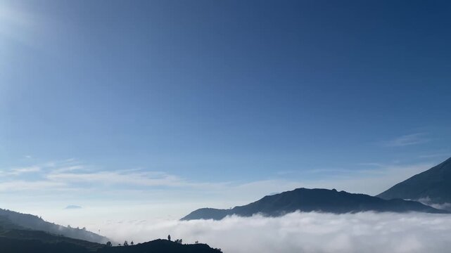 Slow horizontal panning shot capturing a sea of white fog rolling over hills and mountain ridges under a bright blue sky in Central Java, Indonesia.
