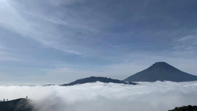 High-angle horizontal panning shot of dense white clouds covering the valley with Mount Sindoro towering in the background under clear blue sky.