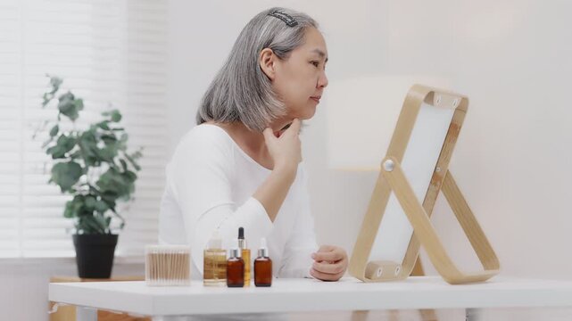 An Asian mature woman with grey hair looks in a mirror with concern while checking fine lines and aging skin.