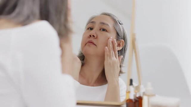An Asian mature woman with grey hair looks in a mirror with concern while checking fine lines and aging skin.