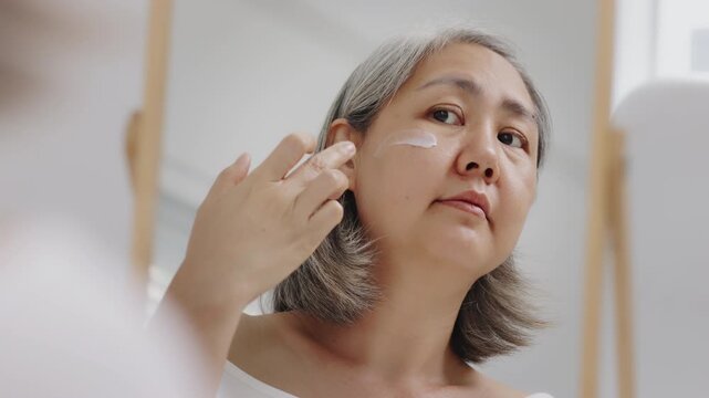 Asian mature woman with natural grey hair applies moisturizing facial cream to her cheek while looking in a mirror. Slow-motion shot.
