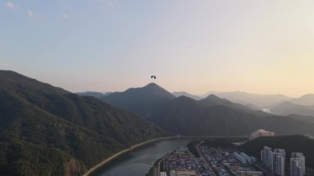 Aerial drone shot orbiting a paraglider gliding over mountain landscape with a town and river view at sunset.
