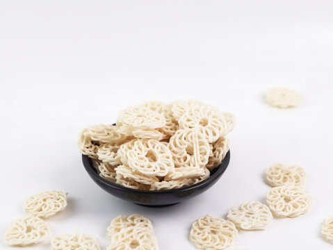 Studio product shot of a black bowl heaped with raw, uncooked white kerupuk udang (traditional Indonesian prawn crackers) alongside loose crackers scattered on a bright white surface.