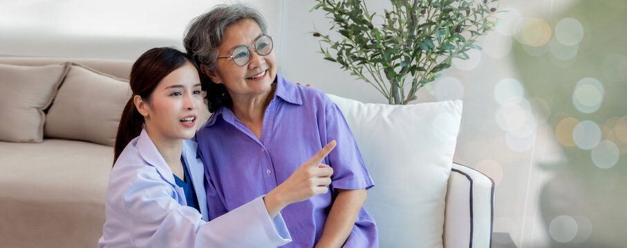Nurse assisting her old patient at nursing home. Caregiver helping senior patient sit on sofa. Friendly female nurse talking with patient at nursing home