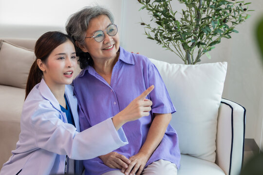 Nurse assisting her old patient at nursing home. Caregiver helping senior patient sit on sofa. Friendly female nurse talking with patient at nursing home