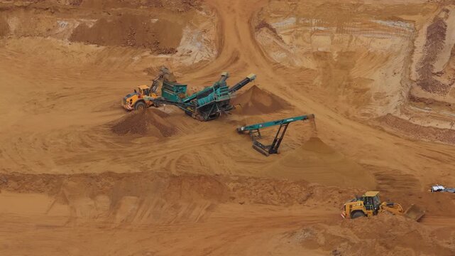 Yellow loaders and industrial conveyor belts move earth in this deep open pit mine during a bright afternoon as workers collect raw material for construction projects across the landscape