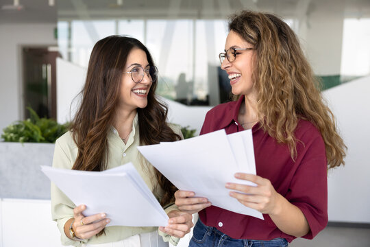 Two smiling businesswomen holding printed documents, reviewing positive report, feeling happy with teamwork results, sharing business success and achievements during collaborative meeting in office