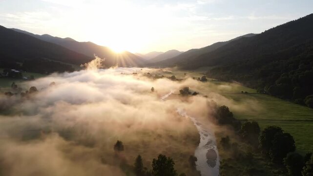 Aerial sunrise view of thick fog rolling across mountain valley floor with river