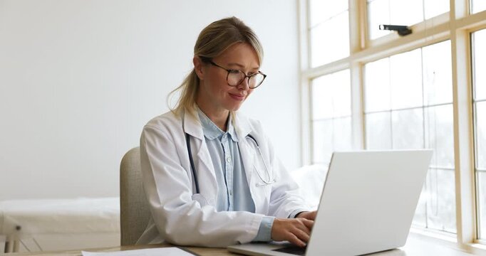 Positive busy Caucasian doctor woman in glasses working on medical reports at computer, analyzing paper records, checking documents, typing on laptop, touching chin, thinking on research