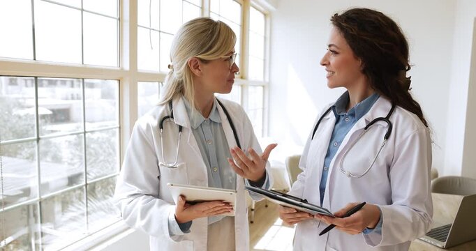 Two positive female doctors talking in clinic office, smiling, discussing medical report, case of patient. Young practitioner woman consulting colleague, asking for advice, mentorship
