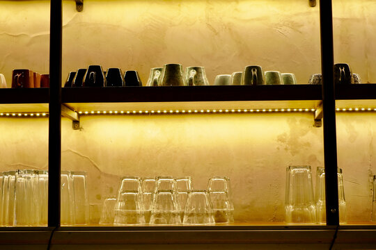 Stacked Glass Cups On Coffee Shop Shelf Inverted glass cups neatly stacked on a wooden display shelf with warm lighting inside a cozy coffee shop, showcasing interior design.