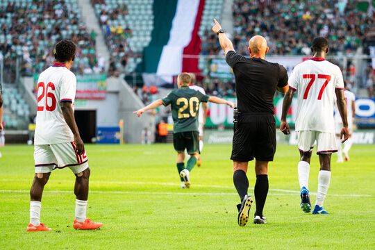 Football referee signaling during a professional soccer match as players react on the field, with stadium spectators creating an intense competitive sports atmosphere.