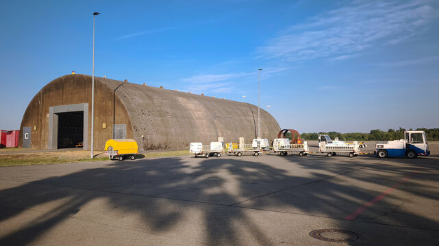 Large concrete arched hangar on an airfield tarmac. Airport tug vehicle towing cargo trailers. Military aviation ground support equipment and storage facility