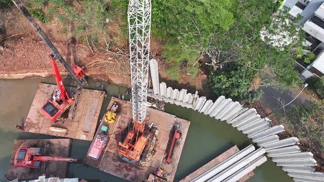 Construction site beside water, surrounded by trees. Orange crane stands tall, supported by wooden planks. White retaining walls line the water's edge. Residential buildings frame the background