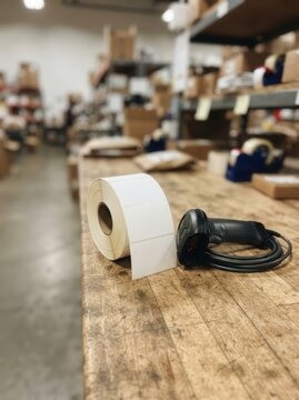 Warehouse scene featuring a roll of blank adhesive labels and a handheld digital barcode scanner on a rustic wooden workbench for logistics