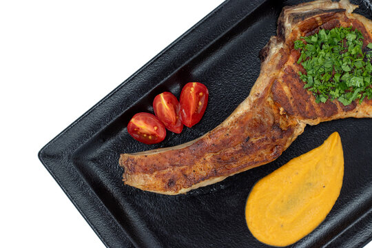 Grilled pork chop with bone served with puree, cherry tomatoes, and fresh herbs on a black plate