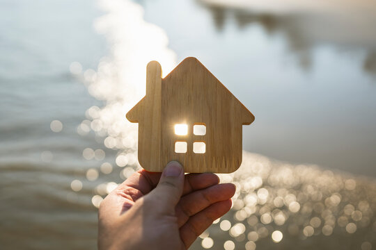 Real estate and dream home concept with hand holding wooden house model silhouette against sunset sunlight on water with glowing bokeh background for property buying and vacation housing.