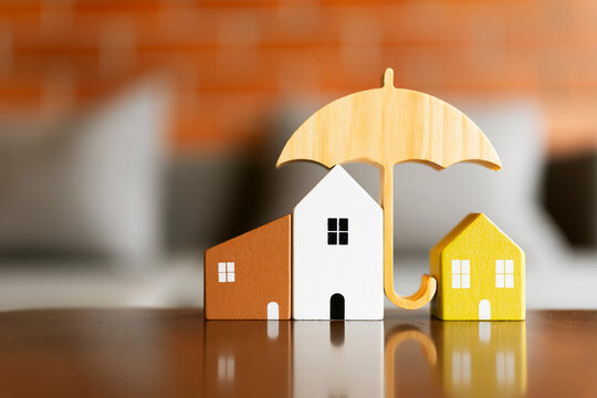 Home insurance and property protection concept with wooden umbrella covering miniature houses on table with warm background for real estate mortgage, safety coverage and risk management.