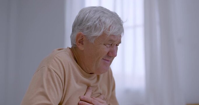 Senior man sits indoors, clutching chest and leaning forward while grimacing from sudden discomfort, expressing visible pain, health concern, vulnerability, and need for immediate caring support.
