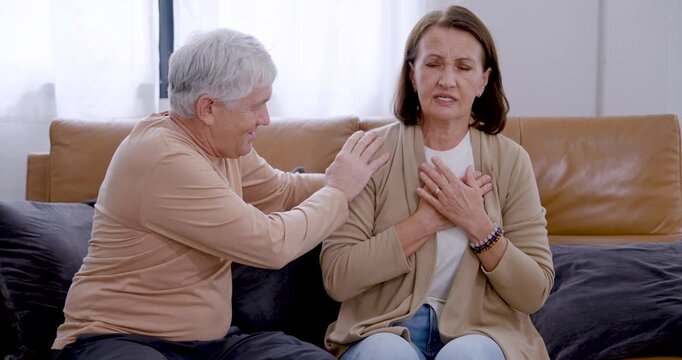 Elderly woman clutches chest and leans forward on living room sofa while supportive husband touches shoulders, providing reassurance, caring attention, comfort, and companionship indoors at home.