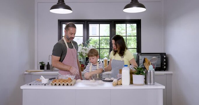 Caucasian mother bends forward to help young son cut and portion dough on kitchen counter while father watches, creating hands on baking lesson with supportive parenting, teamwork and family bonding.