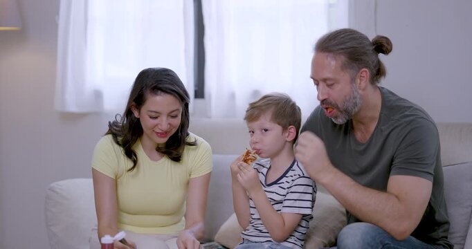 Young son bites snack while Asian mother prepares food and Caucasian father gestures beside him on sofa, sharing family conversation, patient childcare, supportive parenting, and cozy home mealtime.