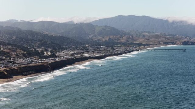 Aerial drone view of the Pacific Ocean coastline, featuring the towns of Daly City and Pacifica with its long pier, nestled against scenic mountains on a clear and sunny day in California
