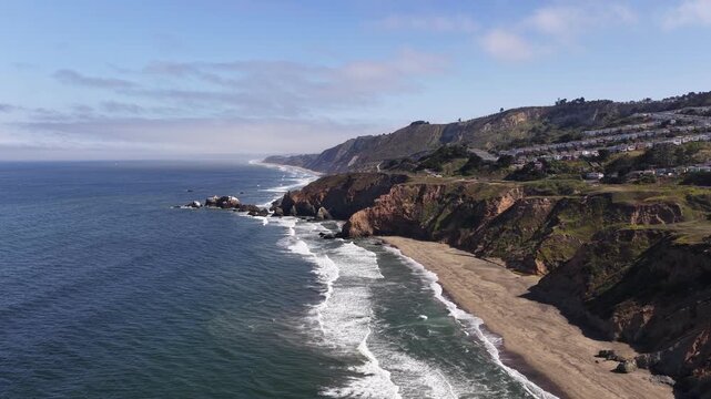 Aerial view captures Mussel Rock Park Beach in Daly City, California, with dramatic ocean cliffs, gentle waves, golden sand, and coastal headlands under clear daylight skies.