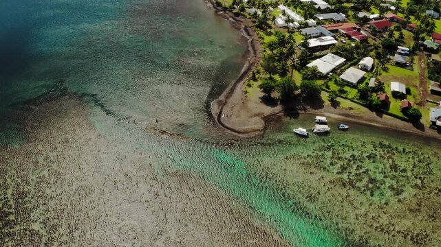 Top down drone shot over textured coral reef patterns near Teahupoo, Tahiti, the edge of a coastal village visible at the corner of frame.